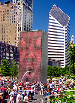 image of 50-foot-tall (15 m) architectural structure spouting water on children from a hole in its face. There is an image of a human face positioned such that the water appears to spout from the mouth. Blue sky and tall buildings are in the background.