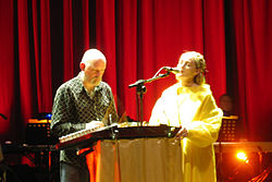 Upper body shot of a forty-five year old man at left. He is peering at a stringed instrument on a lectern. A microphone is directed to a forty-three year old woman at right. She is shown partly in left profile, singing with her eyes closed, her left hand rests on top of the lectern. She wears a ring on her left hand. In the background is musical equipment, a high stage curtain and a foot-light.