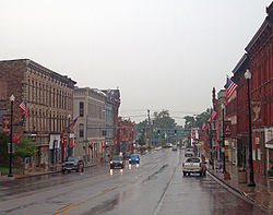 A wide, wet street with tall commercial buildings on either side. Two approaching vehicles have their headlights on, and a traffic light behind them is green. In the rear of the image the street narrows to go over a small bridge.