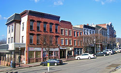 A row of two-story commercial brick buildings, some painted, on a street curving away from the viewer to the right. In front of them are some short bare trees and diagonally parked cars.