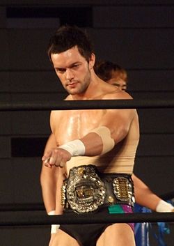 A caucasian male with black hair, and wearing black wrestling tights, standing inside a wrestling ring, with a championship belt around his waist.