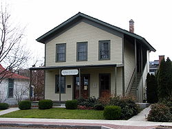 Photograph showing a front view of the Original Wasco County Courthouse, a two-story, wood-frame building with horizontal wooden siding beneath a peaked roof. An exterior stairway leads up the right side of the building, and a covered porch extends across the width of the front of the ground floor. The main doors are centered on the porch and a large sign hangs at the center-front of the porch reading: "Wasco County 1859 / ORIGINAL COURT HOUSE / 401 West Second Place".