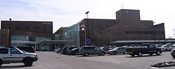 Behind a large parking lot sits an 8-story-tall red brick building with a large number of windows. The words Genesis are on the center of the building at the top. To the left is a slightly shorter building connected to the first with many glass windows. The second building says Pavilion 1