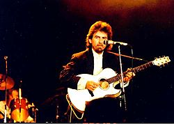 Harrison in his forties, wearing a white shirt and a black jacket, playing a white acoustic guitar and standing behind a microphone. A drummer is partly visible behind him.