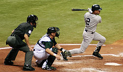 A baseball player in a grey uniform follows through with his swing, while a catcher on the opposing team in a white uniform squats behind him and an umpire in a black uniform stands behind the catcher.