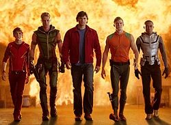 Five men walk away from an exploding building. Each man is dress in an individual costume. From the left, the first man wears an all red outfit with yellow lightning bolts down the side. The second man wears a green, leather outfit. The third man wears a red jacket over a solid blue shirt. The fourth man wears a sleeveless wetsuit, with an orange top and green bottom. The final man wears a silver vest over a black spandex top and bottom.