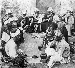 An old photo of a dozen old and middle-aged men sitting on the ground around a mat. A man in front sits next to a mortar and holds a bat, ready for grinding. A man opposite to him holds a long spoon.