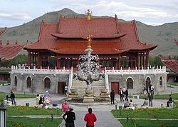 Photograph of a large red-roofed Mongolian building, with an elaborate metallic tree in front of it, about 30 feet tall