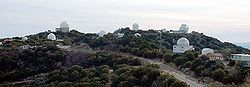 Panoramic view of a mountain top with trees and some white domed telescope buildings and a road leading up to the top.