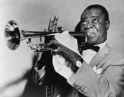 A picture of Louis Armstrong. Short-haired black man in his fifties blowing into a trumpet. He is wearing a light-colored sport coat, a white shirt and a bow tie. He is faced left with his eyes looking upwards. His right hand is fingering the trumpet, with the index finger down and three fingers pointing upwards. The man's left hand is mostly covered with a handkerchief and it has a shining ring on the little finger. He is wearing a wristwatch on the left wrist.