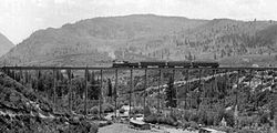 A black and white photograph of a small steam-powered train crossing the bridge, seen from upstream. There is a house along the creek at the bottom
