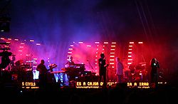 Six musicians playing, from left to right, drums, guitar, keyboards, vocals, bass, and back-up vocals.  They play against a multi-color background with scrolling text by their feet.