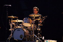 Middle-aged man with slightly graying dark hair and glasses wearing black shirt sits behind a silver-colored, gold-lit drum kit with his left hand and stick about to hit a cymbal and his eyes fixed straight ahead.