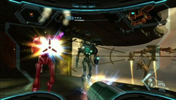 View of a futuristic looking room;  two enemies wearing powered armor are approaching the player, and one is being hit by the player's weapon (a large cannon), which is visible in the corner of the screen. The image is a simulation of the heads-up display of a combat suit's helmet, with a crosshair surrounding the enemy and two-dimensional icons relaying game information around the edge of the frame.