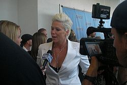 Mia Michaels dressed in a white suit with bleach blond hair standing in front of a journalist while being filmed by an unidentified cameraman.