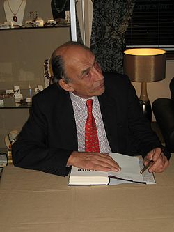 A man wearing a red tie with a chequered shirt and dark jacket signing a book