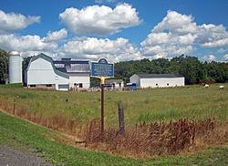 A field with a blue and gold historical marker in the foreground saying "Pottery & Drain Tile Factory Est. ca. 1830 by Fortunatus Gleason & Son Charles, of Morganville, Last Owner was Charles Ford ca. 1900 Excavated by R.M.S.C. 1973. Genesee County Bicentennial". White farm buildings are in the rear, and some cattle are grazing to the right.
