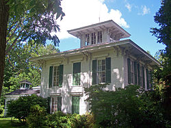 A square white house with green window shutters, wide overhanging eaves and a small square tower in the middle of the roof seen from a corner angle