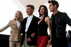 Four people stand beside each other facing off-camera, from left to right: a blonde woman wearing a tan dress suit, a man with salt-and-pepper hair wearing a blazer over white shirt and slacks, a brunette wearing red pants and a black top, and a dark-haired man wearing a black leather jacket over black clothes.