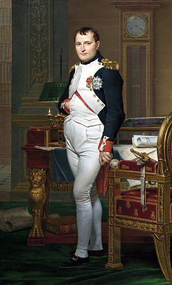 Full length portrait of Napoleon in his forties, in high-ranking white and dark blue military dress uniform. He stands amid rich 18th century furniture laden with papers, and gazes at the viewer. His hair is Brutus style, cropped close but with a short fringe in front, and his right hand is tucked in his waistcoat.