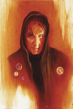 An man with a lined face wearing a hooded jacked and looking into the camera. He wears peace cross, smiley face and fish buttons.