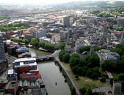 A view from above of office blocks and church spires adjacent to a river which is crossed by a road bridge. In the right foreground a city park and a ruined church. A small boat is moving on the river and a larger barge is moored against a wooded quay. In the distance on the right wooded hills and on the left a mass of predominantly red brick housing.