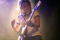 A Caucasian female with shoulder-length brown hair wearing a white shirt and dark pants strums a yellow electric guitar.