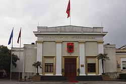 Front of light-colored building with trees, flags and black-on-red Albanian crest