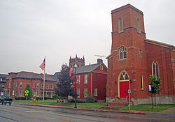 Three brick buildings seen from across a road, slightly to the right. The one at left has a small tower and ornate roofline, the center one is painted red, and the one at right has a square tower in front with a red door.