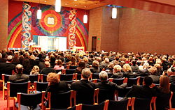 A ground-level view of the backs of around a dozen rows of chairs, filled with men and women, all facing the front of the room. A brightly colored artwork with a rainbow and sun design fills the facing wall. The center of the artwork holds an open Torah ark, and above it is a representation of the two tablets holding the Ten Commandments.