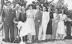 A line of men and women in suits and dresses respectively, standing outside beside a large American flag on a pole. In front of them, a young girl holds a shovel dug into the ground.