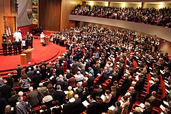 An aerial view of large semi-circular room, filled with rows of curved pews, and a similar balcony on top. People stand in front of most of the seats, holding open books. Many of the men are wearing kippahs (skull-caps). The pews face a raised platform at the front of the room, and in front of that, an open ark containing Torahs. A number of men and women stand in front of the open ark, and above it is a large wall decoration as tall as a person, holding an eternal light.