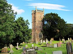Stone building with arched windows and square tower, partially obscured by trees. Gravestones in the foreground