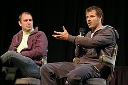 Two seated men. One holds a microphone in one hand and gestures with the other.