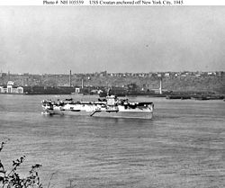 A World War II-era aircraft carrier at anchor. Several aircraft are on her flight deck and land and buildings are visible in the background.