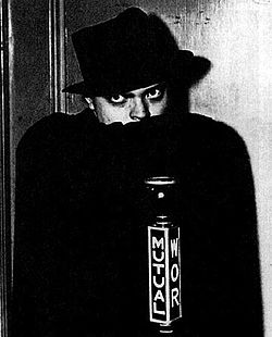 Man in black hat concealing the bottom of his face with a black cape and gazing fiercely. A microphone in front bears the word "Mutual" and the call letters "WOR".