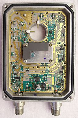 A low-noise block converter with the lid and horn removed exposing the complex circuitry inside, with the exception of the local oscillator which remains covered. The horizontal and vertical polarisation probes can be seen protruding into the circular space where the horn is normally attached. Two output connectors can be seen at the bottom of the device.