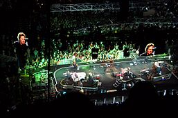 A large arena stage is seen from above with about ten musicians, including a drummer on a stand, as video screens show the singer and green lighting shines on the stage and audience