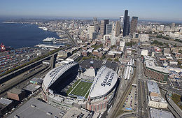 The stadium from the air on a clear day. SEAHAWKS STADIUM is painted on the white partial roof. The stadium is surrounded by roads and buildings.