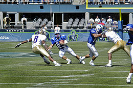 In an American football game, a runner with the ball faces a defender while a blocker locks with another defensive player.