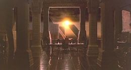 A screen-shot from the film depicts a large room as we look between two rows of pillars. At the far end of room there is a massive window that dwarfs a man in front of it. The man is facing away from us looking out of the window, through which the sun can be seen.