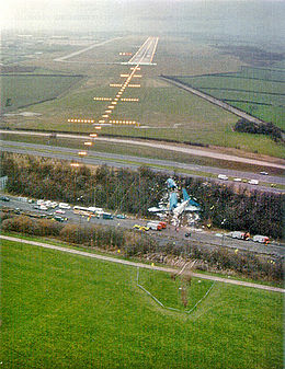The wreck of an airliner lies between roads roughly 100m to the right of approach lights and several hundred metres in front of a runway. The wreck is broken into three large pieces, a nose section, a central section and a tail section. The tail section is turned around, the horizontal stabilizers resting in front of the wings of the central section.