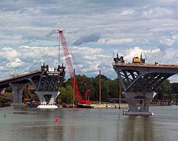 Two level metal structures on concrete supports on either side of a river. There is construction equipment on both of them and a large red crane on the far shore