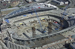 A stadium under construction with two cranes positioned where the field will eventually be installed. The terraced seating sections rise above the partially complete concourse levels, and half of the roof is in place.