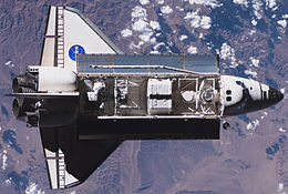 A space shuttle, with its payload bay full of equipment, seen orbiting over a cloudy sky above a mountainous region of the Earth.