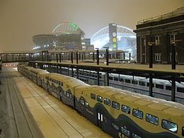 The last commuter train at a train station with a brightly lit stadium nearby. The stadium's roof supports are colored with green and red lights for the Christmas season.