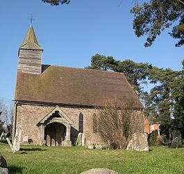 A stone church with a tiled roof seen from the south. On the left gable is a shingled bellcote, and a wooden porch protrudes from the church