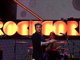 A thirty-six year-old man is shown in three-quarter view. He is standing centre-stage, half-turned with hands clapped together. He is partly obscured by a microphone stand in front. Behind him is a drummer at his kit. Beyond that is the lighting, which includes the partly spelled letters R-O-C-K-C-O-R in large orange-yellow.