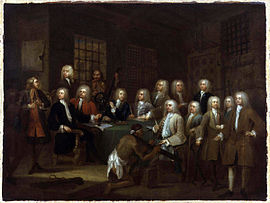A painting of six men wearing white wigs, and dressed elegantly, seated around a table covered with a green cloth. Eight men, also wearing wigs and dressed in finery, are standing behind the table. One man without a wig, dressed in a rougher style, and with a rough-looking face, stands at the front of the table, addressing the others. Another rough-looking man stands at the back. A black man wearing only a cloth around his waist kneels in front of one of the men wearing a wig. The black man is wearing an iron device around his neck. Another iron device is lying on the floor behind him.