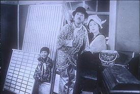 A young girl, man, and woman standing outside of a house, all looking up in the sky. The girl, on the left, is smiling and pointing skyward. The man wears a bowler hat and holds a short broom over his shoulder; the woman wears a kerchief around her head. They are surrounded by domestic objects as if just moving into or out of the house.